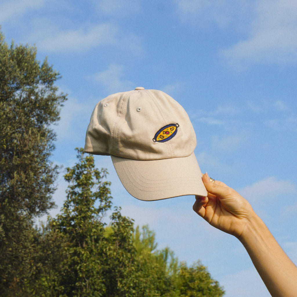 Beige cap with a paella design held against a blue sky with trees
