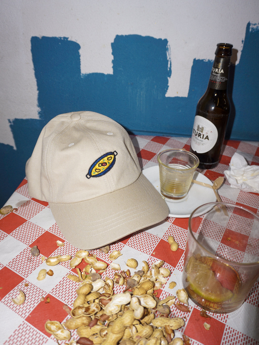Beige cap, bottle, glass, and peanuts on a checkered tablecloth with blue wall background