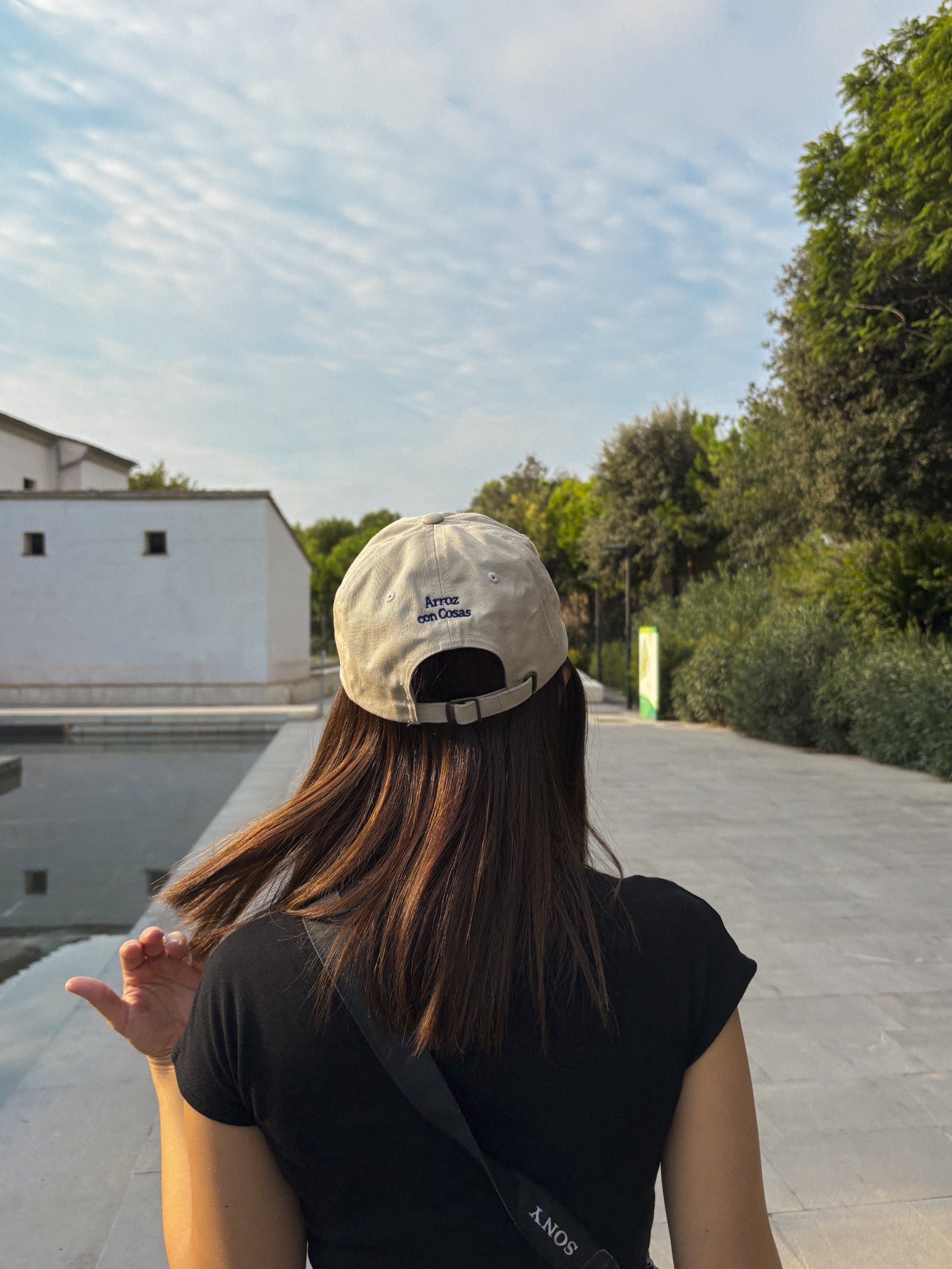 Person wearing a beige cap, walking outdoors with trees and building in the background